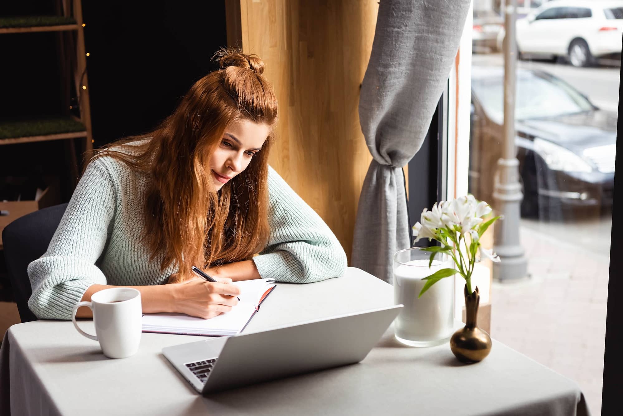 redhead woman writing in notepad while watching webinar on laptop in cafe